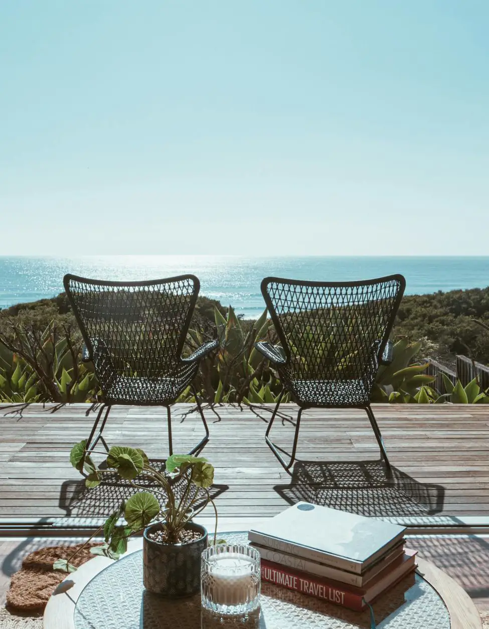 Two black wicker chairs face an ocean view on a wooden deck under a bright blue sky. A small table with a plant, candle, and stacked books titled 'Ultimate Travel List' is in the foreground. Lush green foliage extends to the horizon where the ocean meets the sky.
